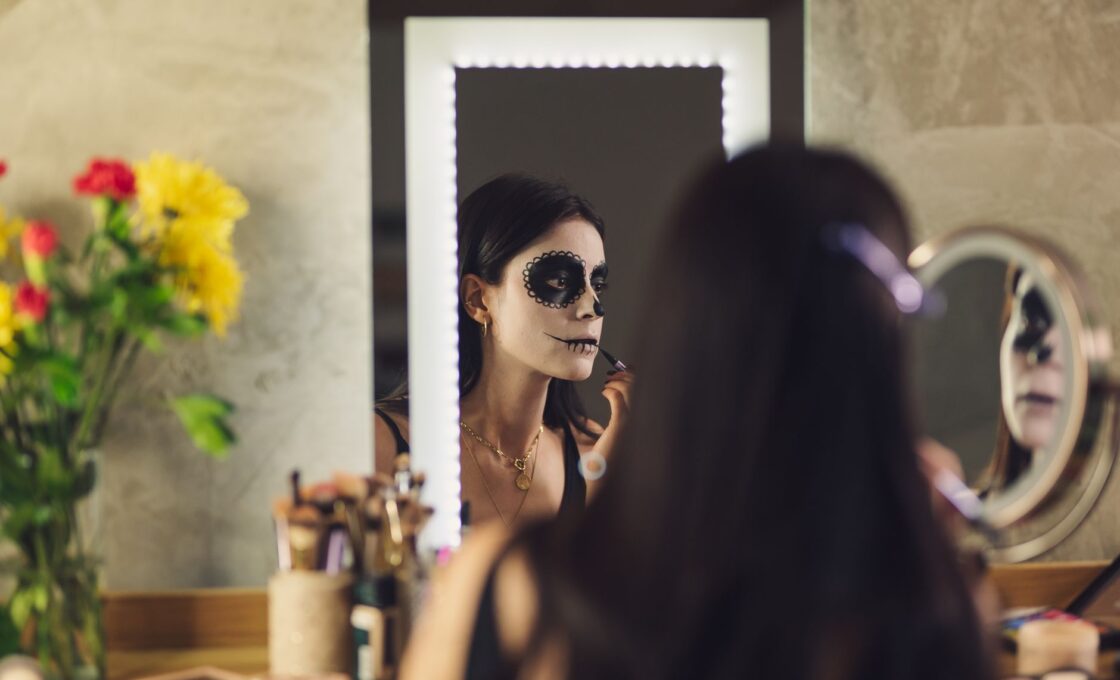 Woman applying skull face paint in front of a mirror, a reminder to keep costume makeup away from the eye area
