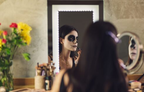 Woman applying skull face paint in front of a mirror, a reminder to keep costume makeup away from the eye area