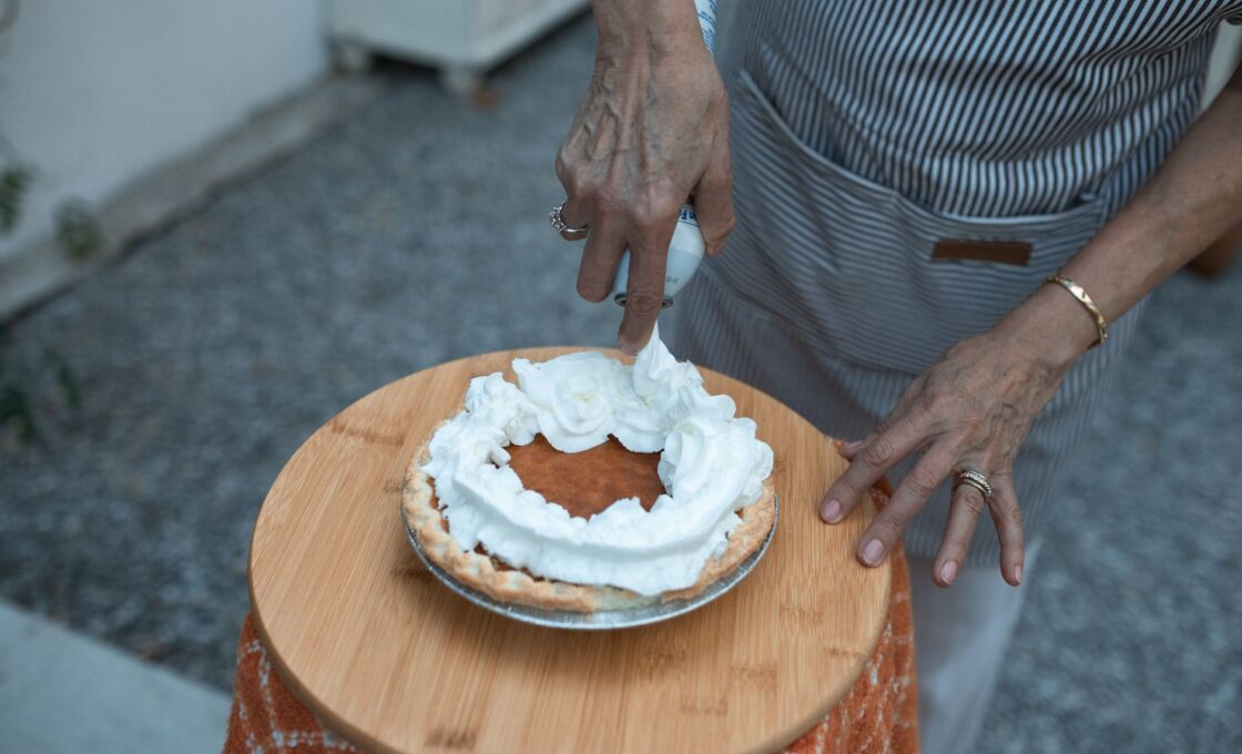 Hands adding whipped cream to a slice-ready homemade pumpkin pie for Thanksgiving
