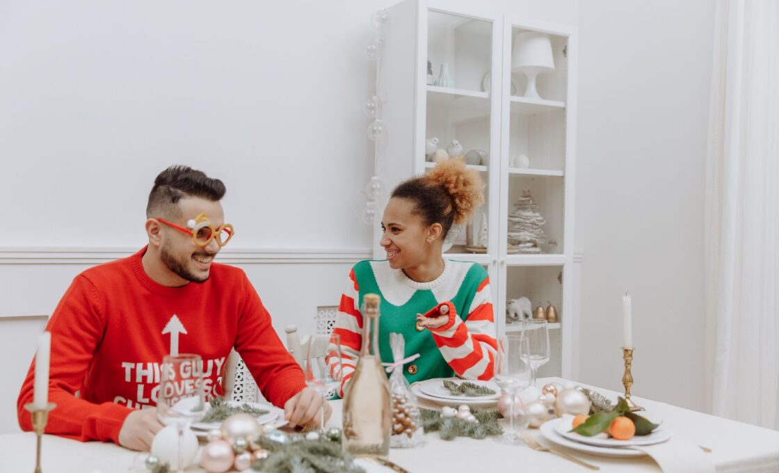 Couple in holiday sweaters sitting at a festive dinner table, planning how to split the holidays between families