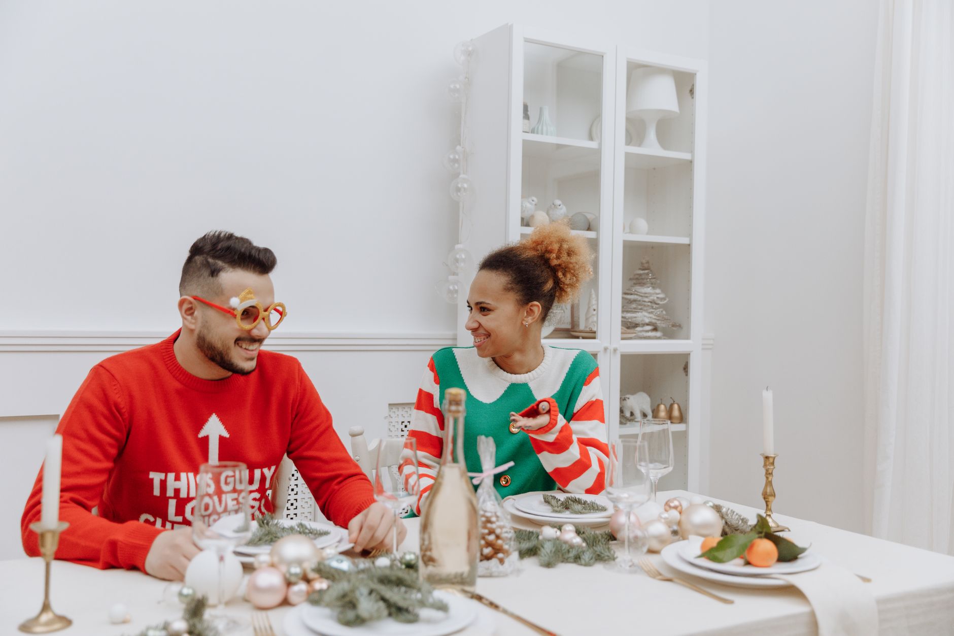 Couple in holiday sweaters sitting at a festive dinner table, planning how to split the holidays between families